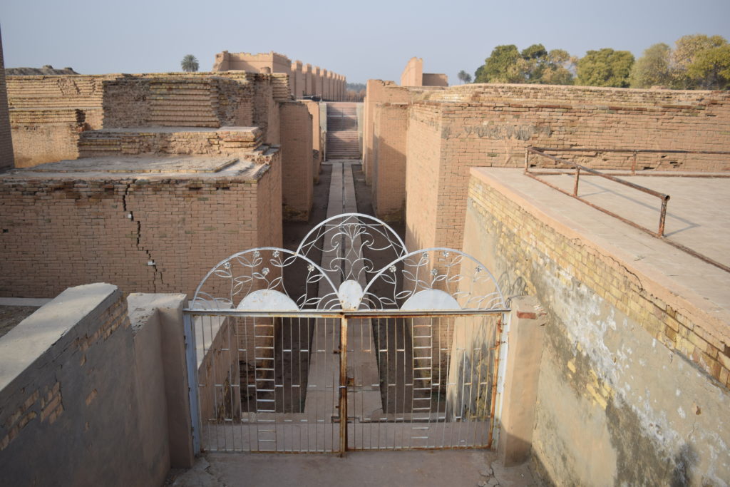 image illustrating the archeological ruins of babylon in iraq. a modern gate appears in the foreground, with remains of brick structures in the background