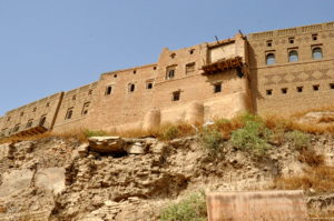 image illustrating the external wall of the heritage citadel of erbil in the Kurdistan Region of Iraq. an imposing brick wall appears above with damage to a balcony visible